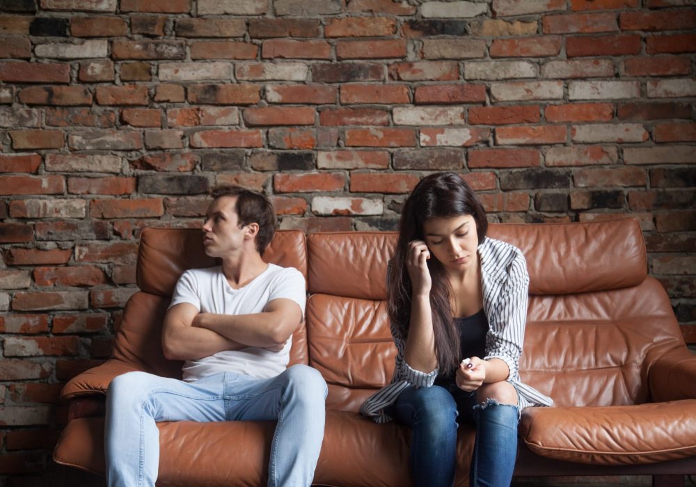Frustrated upset couple in quarrel not talking after fight, offended stubborn insulted jealous man sitting silent on sofa at home with sad depressed disappointed woman, marriage relationships problem