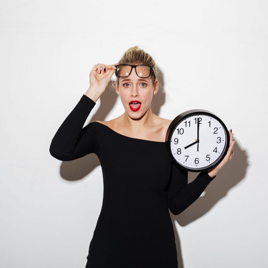 Photo of young shocked business woman standing isolated over white background. Looking camera holding clock.